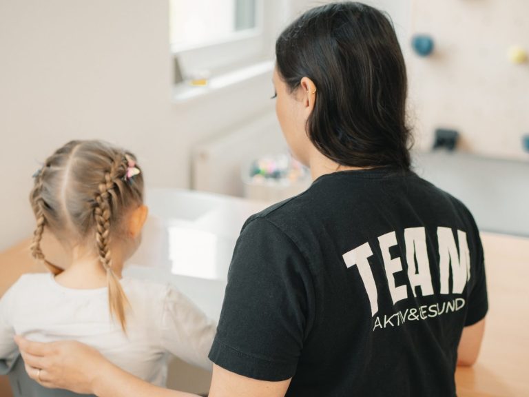 Ein Erwachsener steht hinter einem Kind, beide schauen auf einen Tisch in einem hellen Raum.