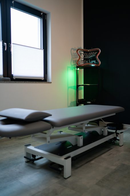 Behandlungsliege in einem Raum mit Fenster und dekorativem Regal im Hintergrund.